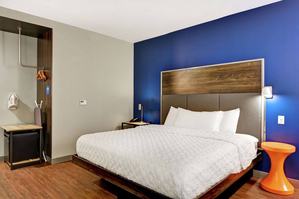 A modern hotel room with a large bed, a wooden headboard, a nightstand, a small table, and a unique orange stool.
