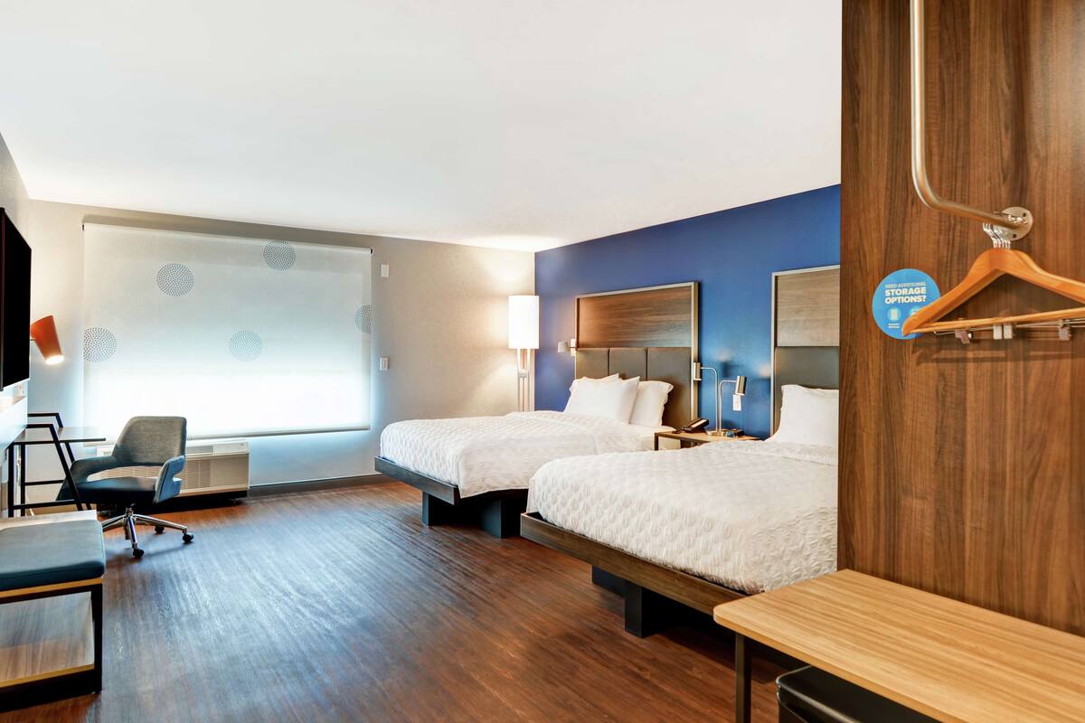 A hotel room with two beds, a desk, a chair, and a large window with blinds.