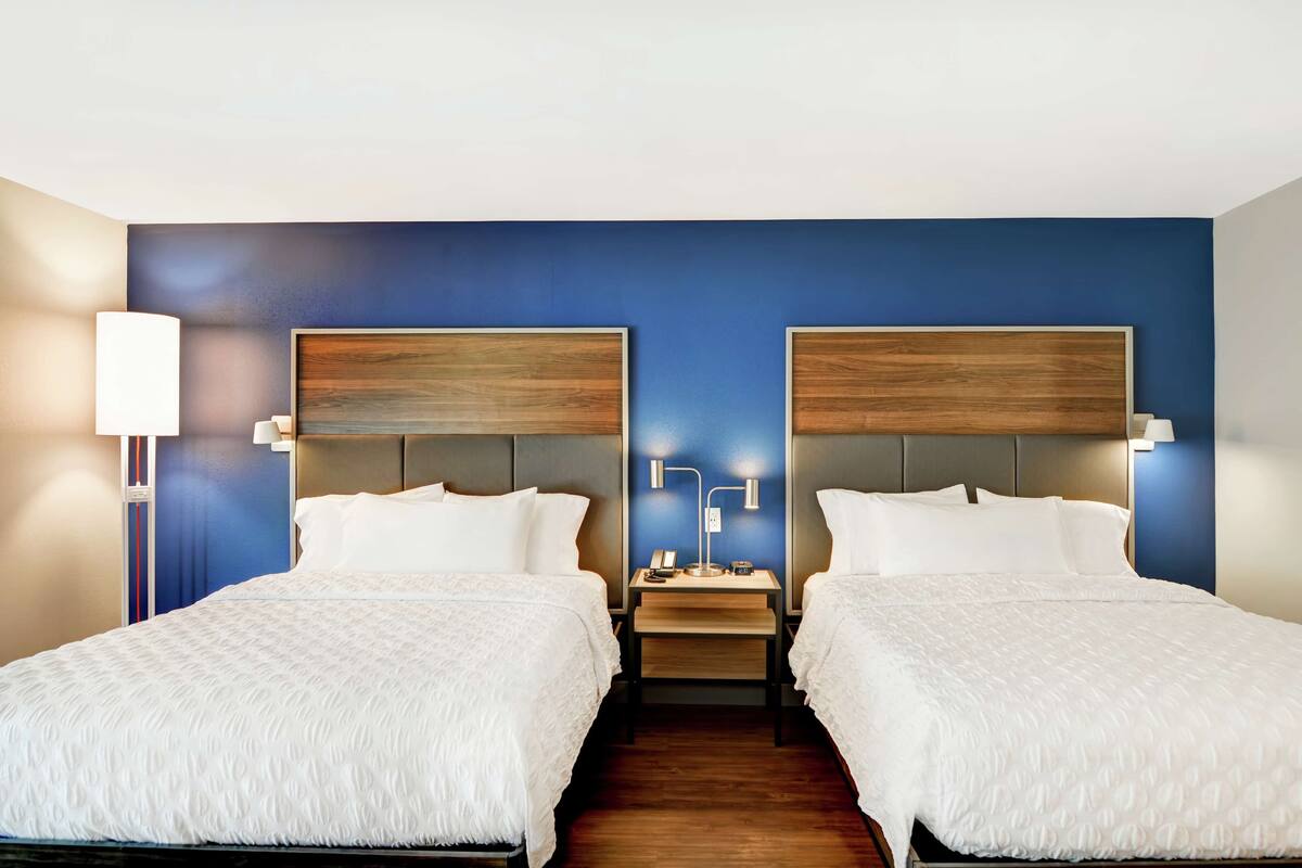 A hotel room with two beds, wooden headboards, bedside tables with lamps, and a blue accent wall.
