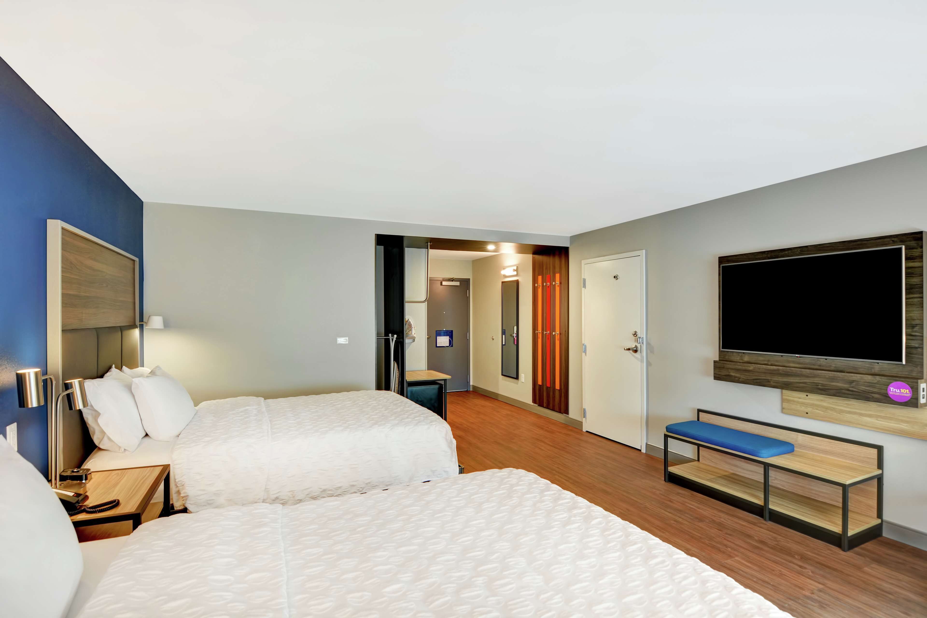 A hotel room with two beds, a bench, a TV, and a door leading to a bathroom.