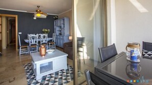A modern living room with a glass dining table, a ceiling fan, and a kitchen area with cabinets and a chair. - NAIMA Apartment in Calpe – 1st line on Fosa Beach, for 4 people, terrace, swimming pool, Wi-Fi and air conditioning (Calpe)