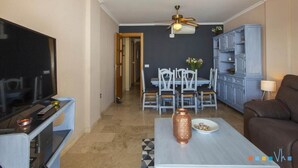 A living room with a flat-screen TV, a coffee table with a copper vase, a brown sofa, and a dining area with a table and chairs. - NAIMA Apartment in Calpe – 1st line on Fosa Beach, for 4 people, terrace, swimming pool, Wi-Fi and air conditioning (Calpe)