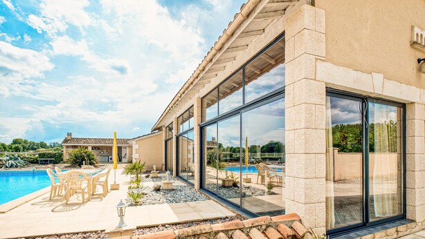 Een modern gebouw met grote glasramen, een zwembad, terraszitplaatsen en een helder uitzicht op het omliggende landschap.