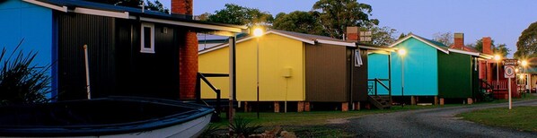 A row of colorful, single-story buildings with a gravel path leading to a sign indicating a speed limit of 8. - Lake Conjola Waterfront Holiday Park (Lake Conjola)