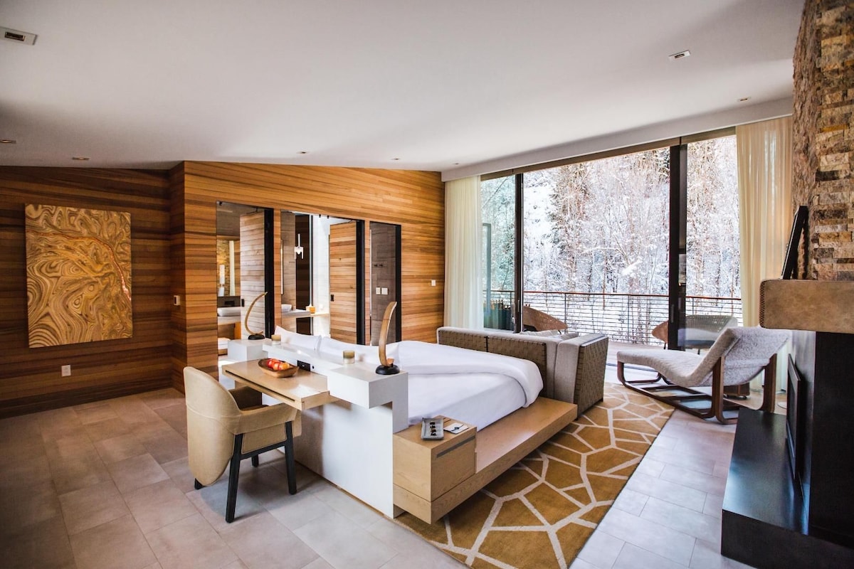 Una habitación de hotel moderna con una cama grande, un escritorio, una silla, un espejo amplio y un balcón con vistas a árboles.