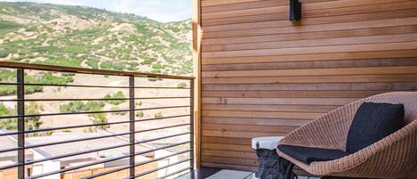 A balcony with a wicker chair and a small trash bin, overlooking a hilly landscape.