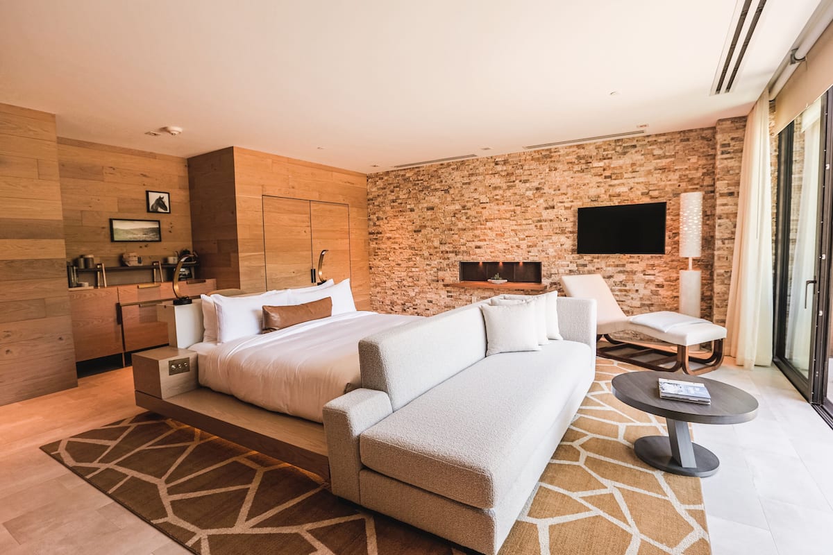 A modern hotel room with a large bed, a sofa, a dining table, a TV, and a fireplace.