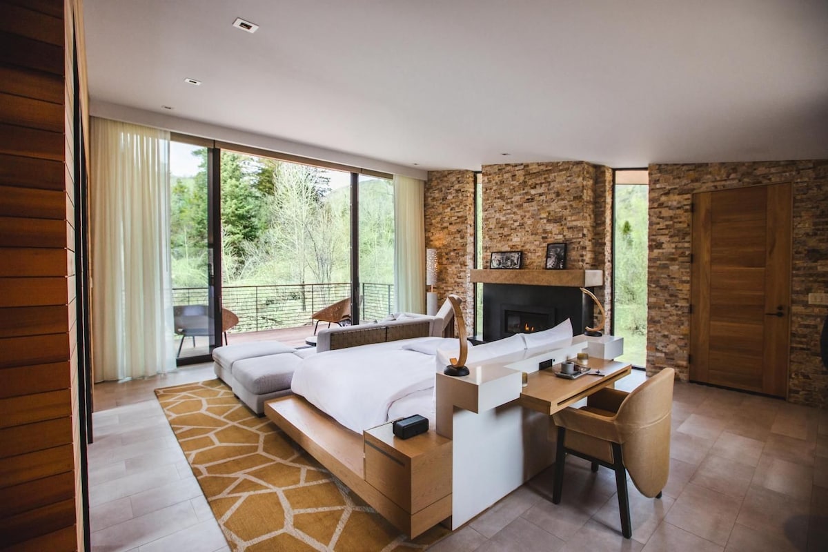 Un dormitorio moderno con cama, escritorio y chimenea, ventanas amplias y un balcón.