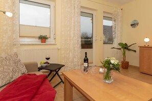 A cozy living room with a sofa, a dining table with a bottle and glasses, a potted plant, and a window with curtains.