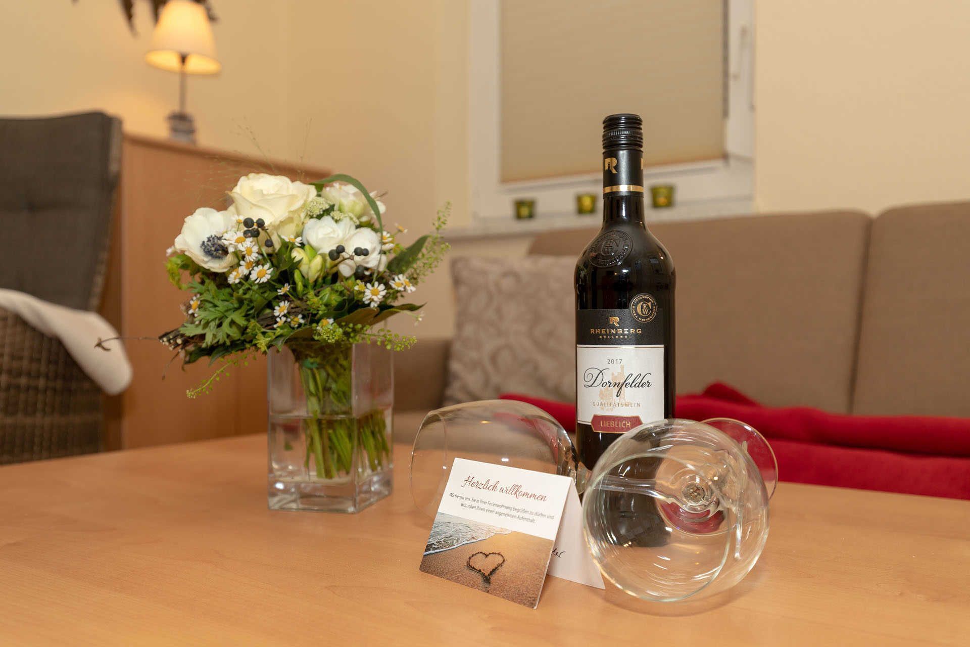 A table with a bottle of wine, a glass, a card, and a vase of flowers.
