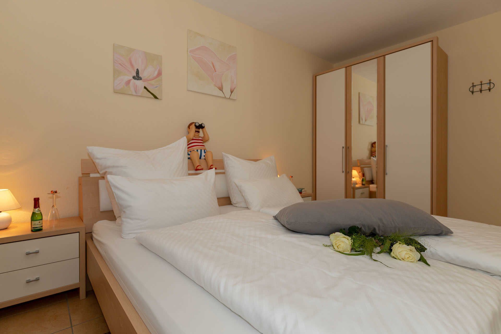 A bedroom with a bed, pillows, a nightstand with a lamp and bottle, a dresser, and a wall with two flower paintings.