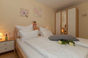 Ein Schlafzimmer mit einem Bett, Kissen, einem Nachttisch mit Lampe und Flasche, einer Kommode und einer Wand mit zwei Blumenbildern.