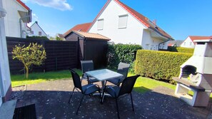 Eine Terrasse mit Tisch und Stühlen, ein kleiner Baum, ein Hecke und ein Haus mit rotem Dach im Hintergrund.
