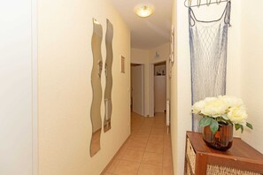 Ein Flur mit Fliesenboden, einem Spiegel an der Wand, einer Tür am Ende und einer Vase mit Blumen auf einem Tisch.