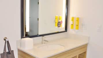 Un baño con un espejo grande, un lavabo y un mueble de tocador de madera. Hay artículos de aseo en la pared y un bolso marrón colgando.
