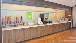 Een ontbijtbuffet met yoghurt- en havermoutautomaten, een koffieapparaat en een bord dat 'Top T Breakfast' aanprijst.
