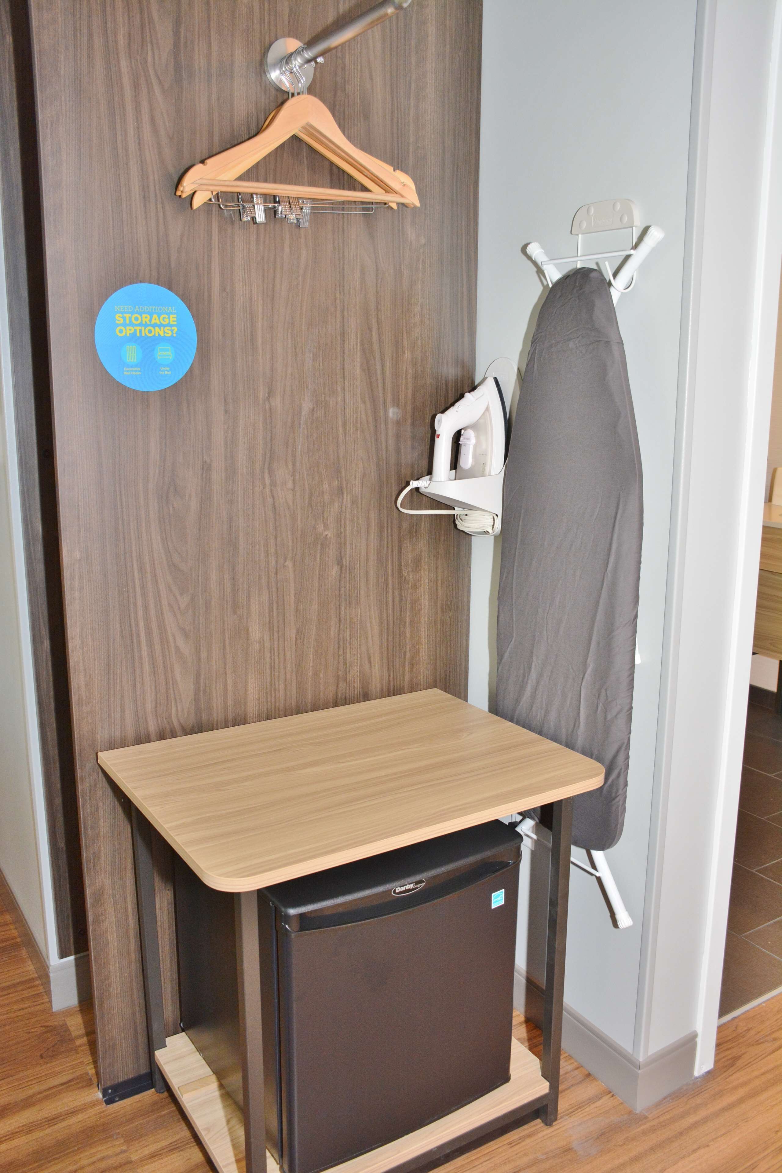 Una habitación pequeña con una mesa de madera, un refrigerador compacto, un perchero y una tabla de planchar fijada a la pared.