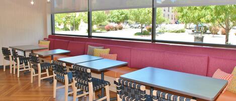 A restaurant with wooden chairs and tables, large windows with a view of the street, and a red cushioned bench area.