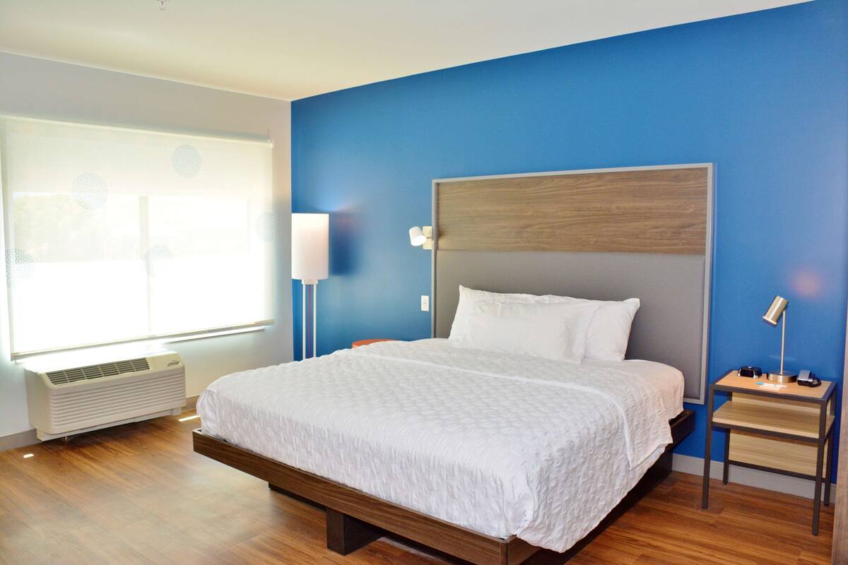 Een hotelkamer met een groot bed, een nachtkastje met een lamp, een raam met zonwering en een wandairconditioner.