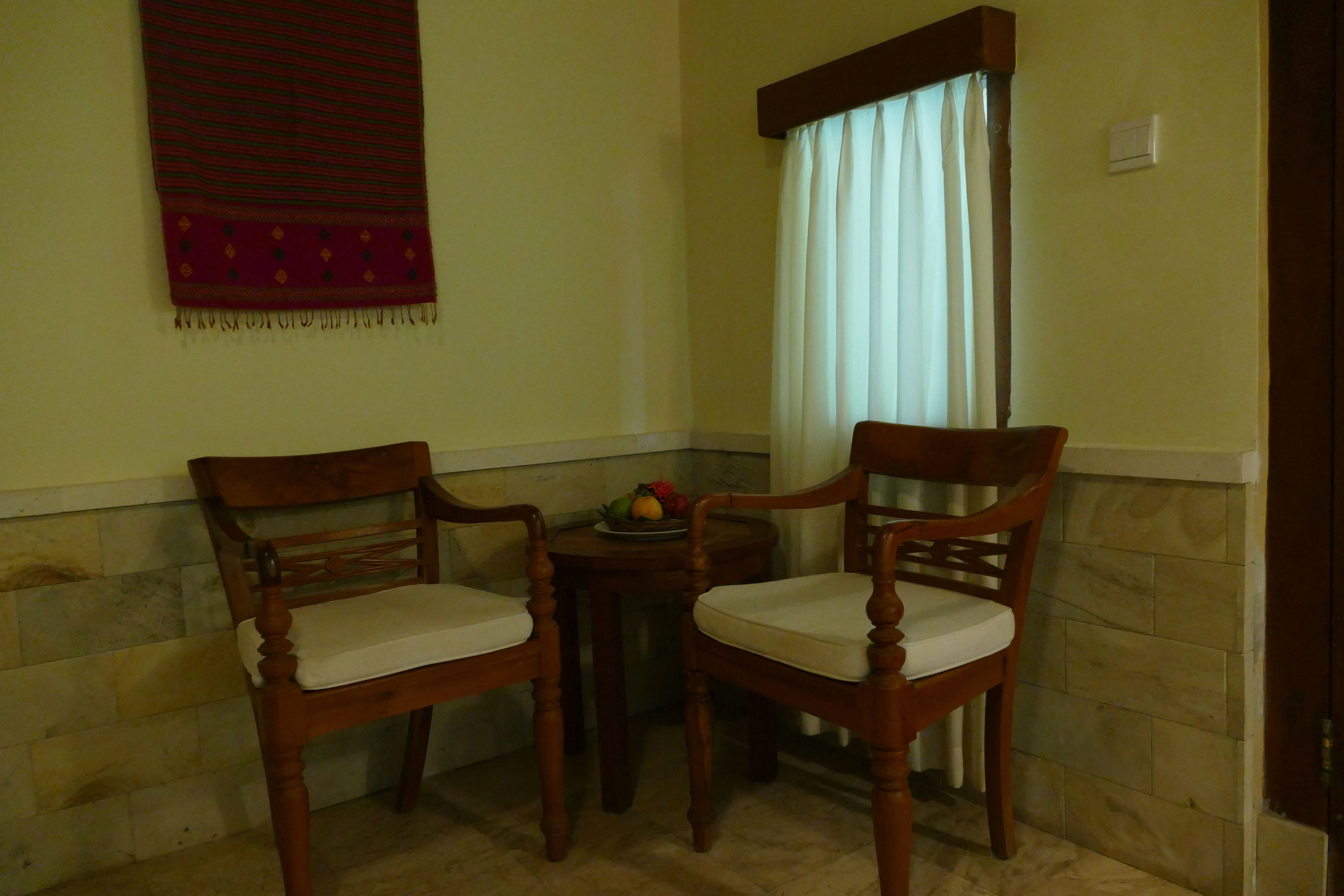 Two wooden chairs with white cushions, a small table with a fruit bowl, and a window with curtains.