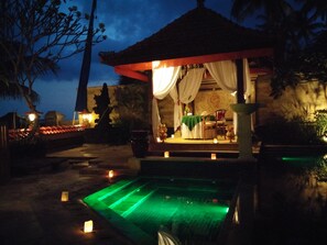 Una glorieta con un comedor, que incluye una mesa y sillas, rodeada de velas y una zona de piscina iluminada en verde.