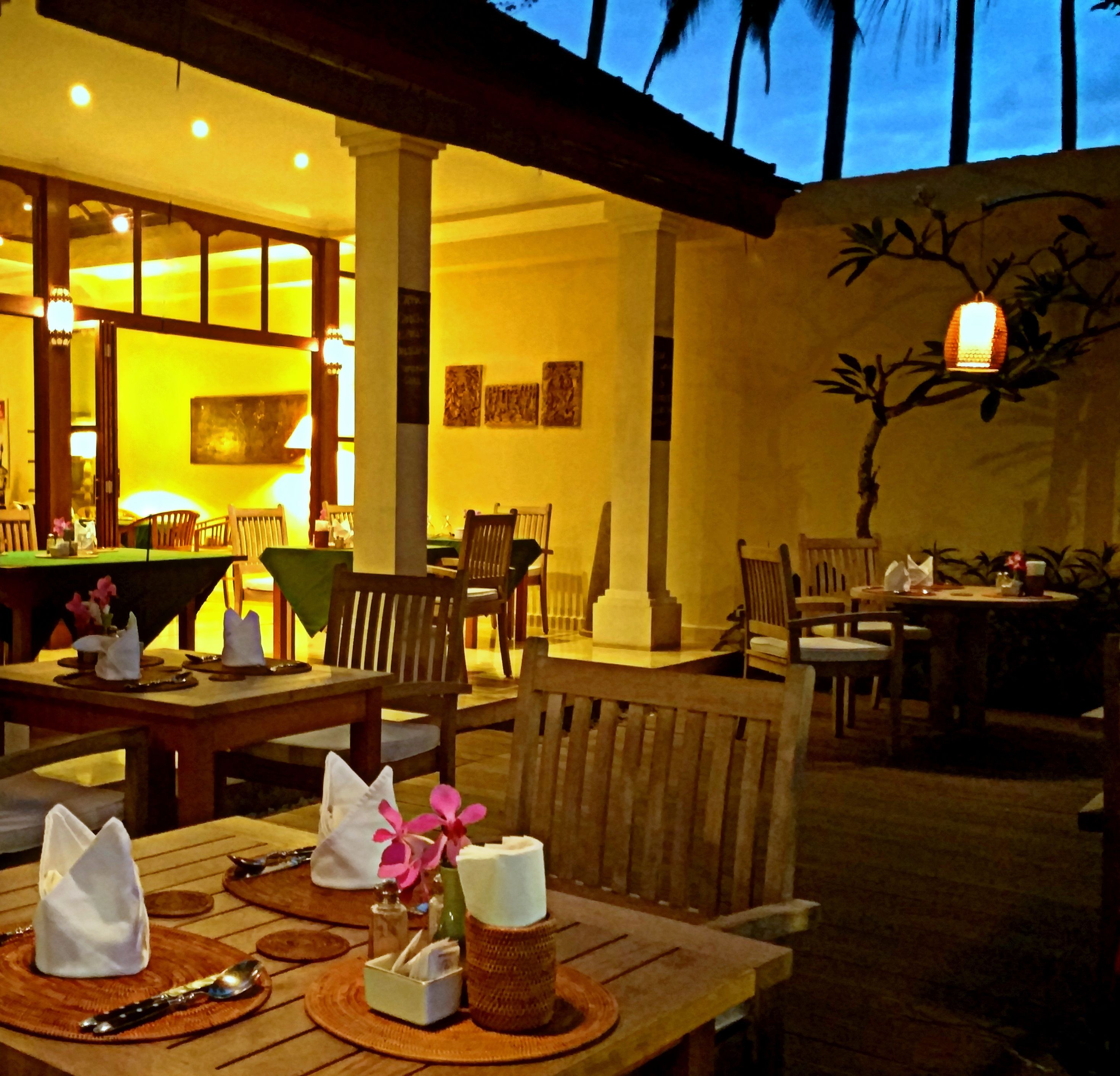 Un espacio exterior para comer con mesas y sillas de madera, un árbol decorativo e iluminación ambiental cálida.