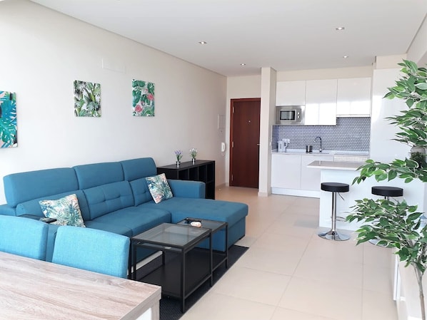 A modern living room with a blue sofa, a small coffee table, and a dining table. There is a kitchen area with white cabinets and a microwave.