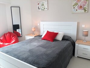 A modern bedroom with a large bed, a red bean bag chair, two bedside tables with lamps, and floral artwork on the walls.