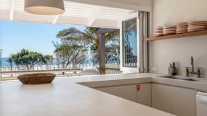 Eine moderne Küche mit einem großen Fenster mit Blick auf einen Strand, eine Holzschüssel auf der Arbeitsplatte und ein Regal mit Tellern.