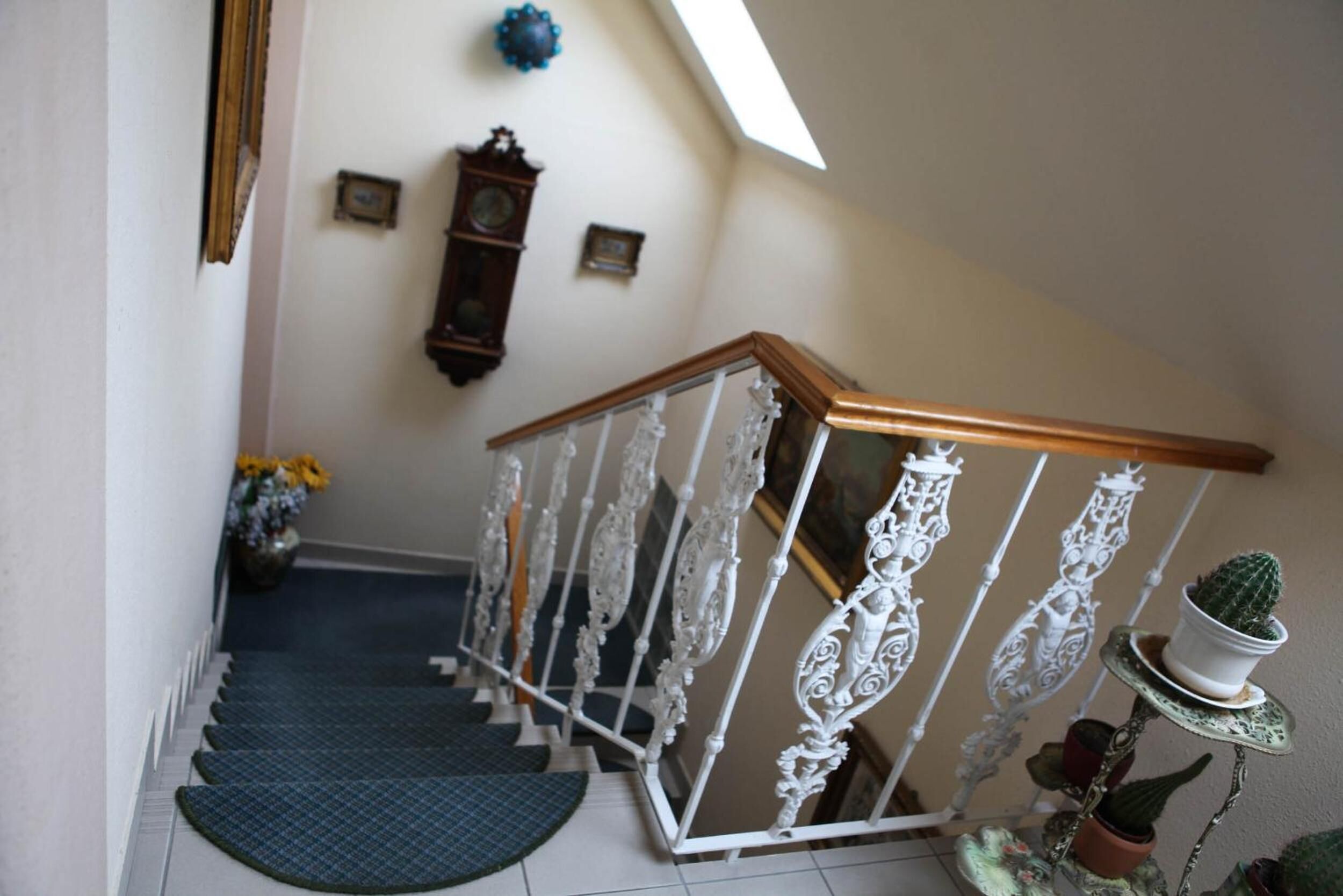 Un escalier orné de balustrades blanches et doté d’une main courante en bois, menant à un palier où se trouvent une horloge et des tableaux encadrés accrochés au mur.