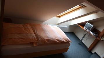 Ein Einzelbett mit orangefarbener Bettdecke in einem Zimmer mit Oberlicht, einem Schreibtisch mit Computer und einem Stuhl.