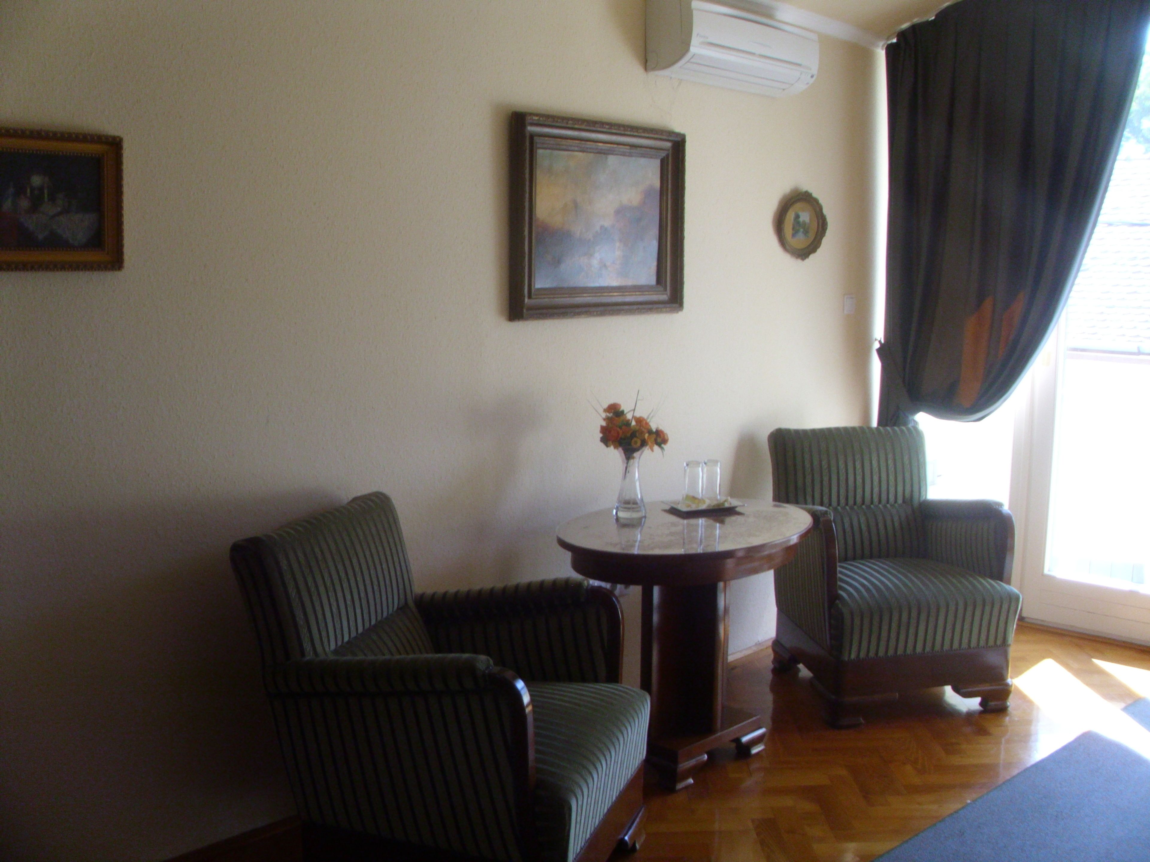 A room with two upholstered chairs, a small round table with a vase and glasses, a framed picture on the wall, and a window with curtains.