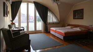 Un dormitorio con cama, una silla, una mesa con un florero y vistas al exterior a través de un ventanal con cortinas.