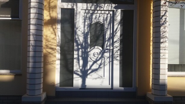 Una puerta principal con un panel de vidrio y una mirilla, flanqueada por dos columnas, con sombras de árboles sobre el vidrio.