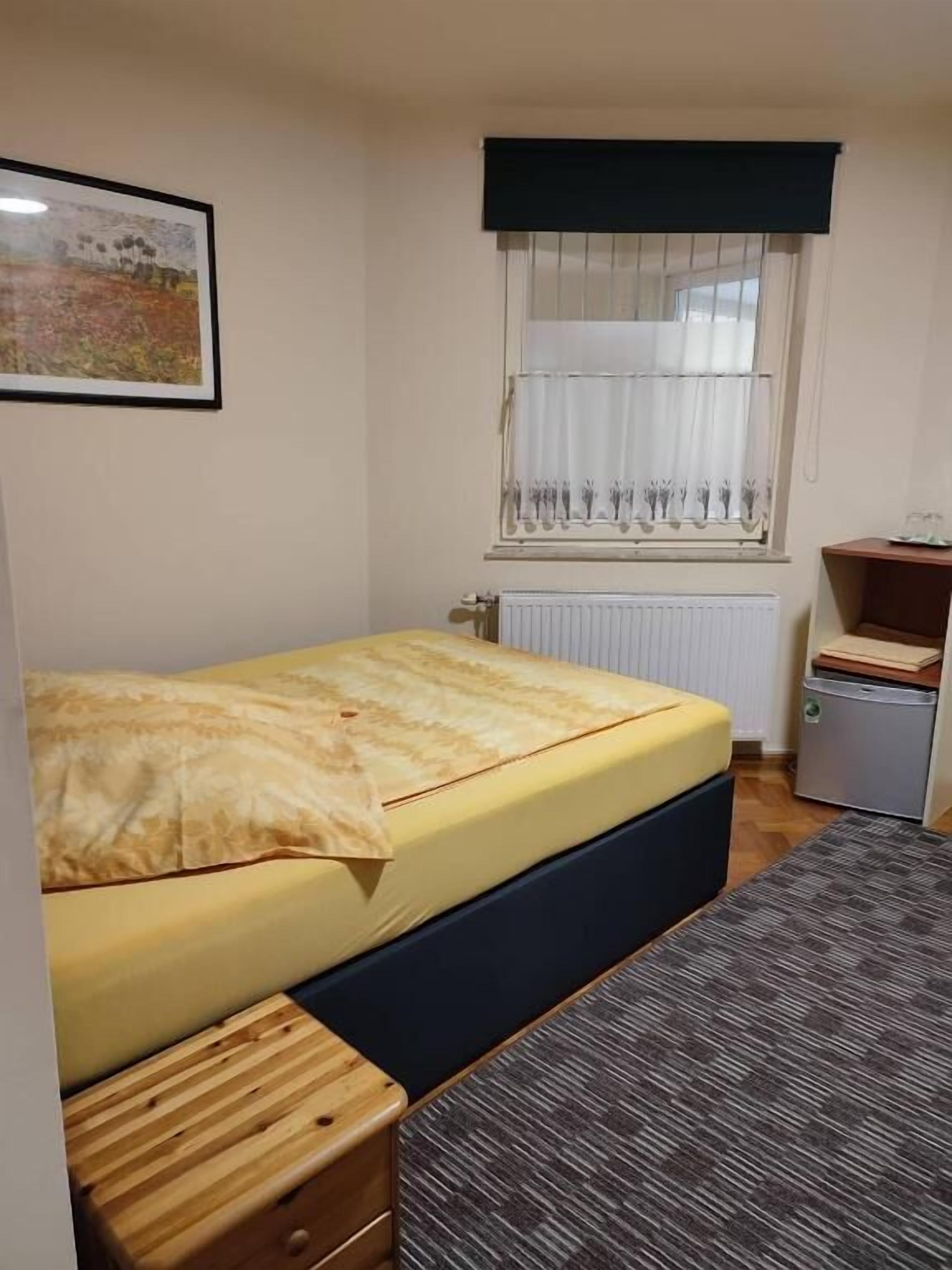 Un dormitorio pequeño y ordenado con una cama individual, una mesita de madera, un radiador, una ventana con cortinas y un cuadro en la pared.