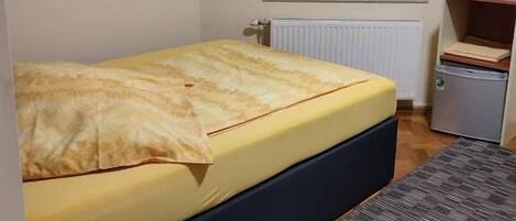 Un dormitorio pequeño y ordenado con una cama individual, una mesita de madera, un radiador, una ventana con cortinas y un cuadro en la pared.