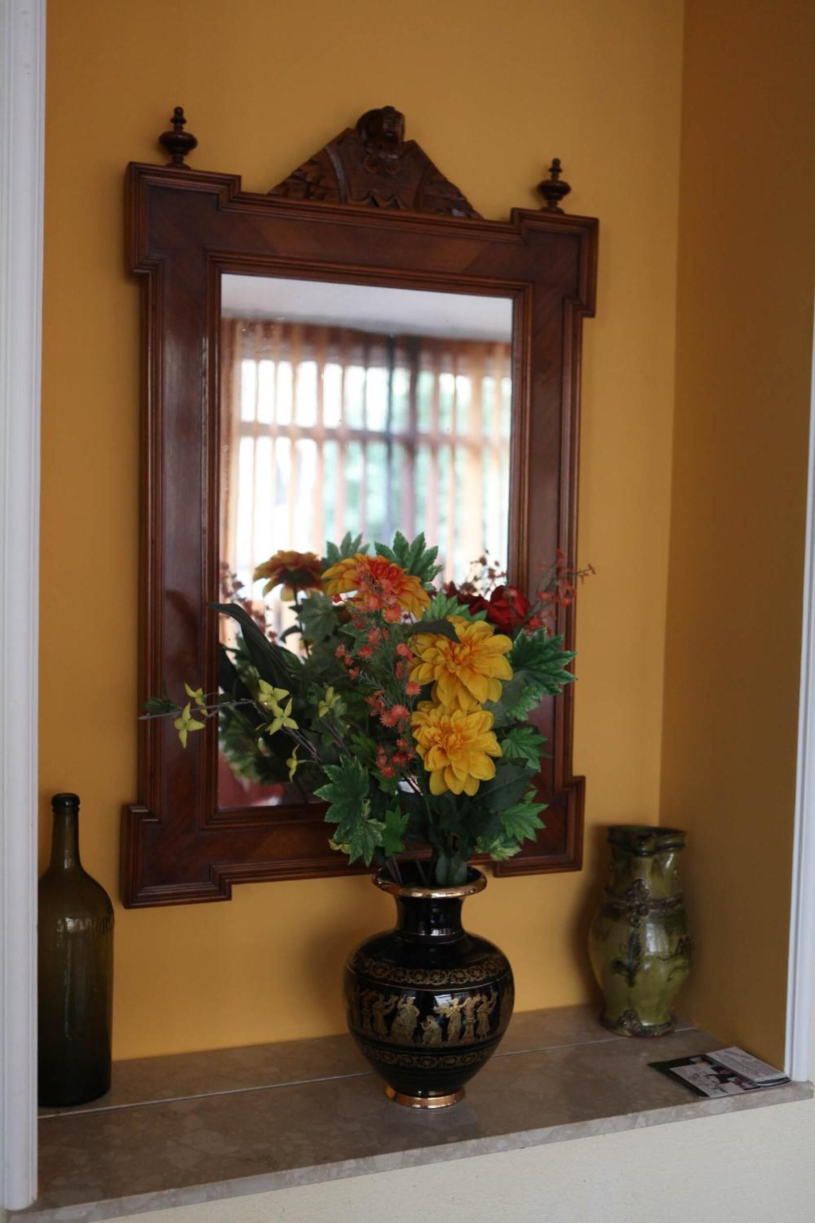 A mirror with a wooden frame, a vase with flowers, and a bottle on a shelf.