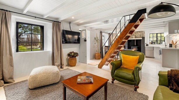 A spacious living area with a wooden staircase, a green armchair, and a modern kitchen in the background. - Wanaka Stone Cottage - Wanaka Holiday Home (Wanaka)