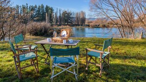 A picnic setup with a table and chairs by a lake, surrounded by trees and a clear sky. - Wanaka Stone Cottage - Wanaka Holiday Home (Wanaka)