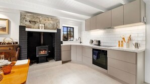 A modern kitchen with a stone fireplace, white cabinetry, and a dining area with a table and chairs. - Wanaka Stone Cottage - Wanaka Holiday Home (Wanaka)