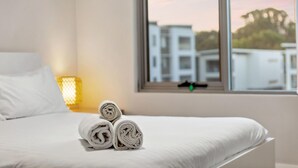 A neatly made bed with rolled towels, a window with a view of buildings, and a lamp on the nightstand. - Rockingham Relaxed Apt | Fast Wi-fi + Free Parking - 2 Bed 2 Bath Standard (Rockingham)