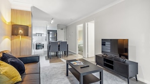 A modern living room with a sofa, coffee table, TV stand, and kitchen in the background. - Rockingham Relaxed Apt | Fast Wi-fi + Free Parking - 2 Bed 2 Bath Standard (Rockingham)