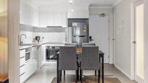 A modern kitchen with white cabinetry, a built-in oven, a refrigerator, and a dining area with a table and chairs. - Rockingham Relaxed Apt | Fast Wi-fi + Free Parking - 2 Bed 2 Bath Standard (Rockingham)