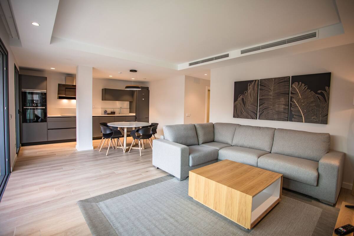 Una sala moderna con un sofá seccional gris, una mesa de centro de madera y un comedor con una mesa redonda y sillas.