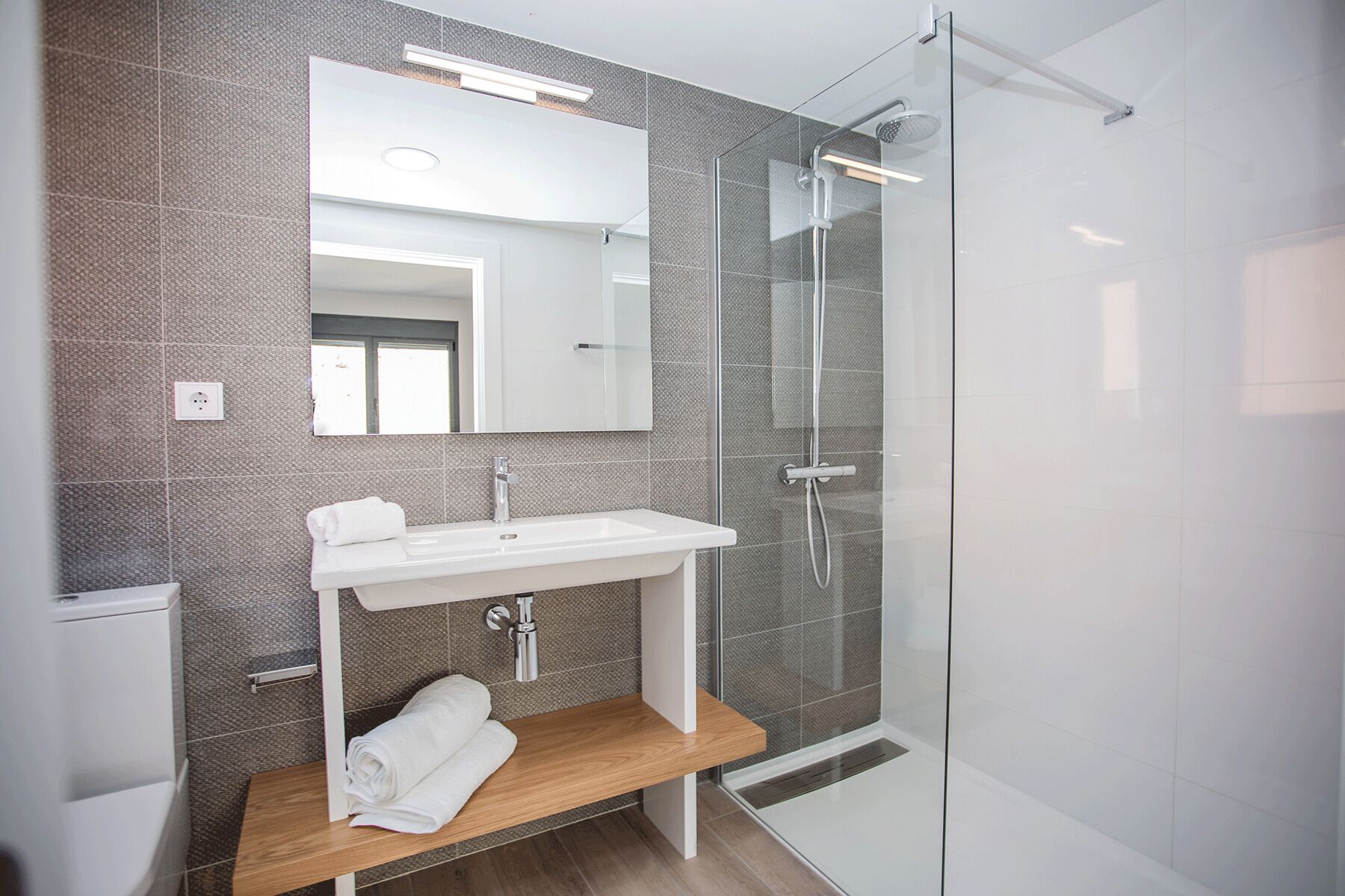 Une salle de bain moderne avec une douche à parois de verre, un lavabo blanc et une étagère en bois où sont posées des serviettes.
