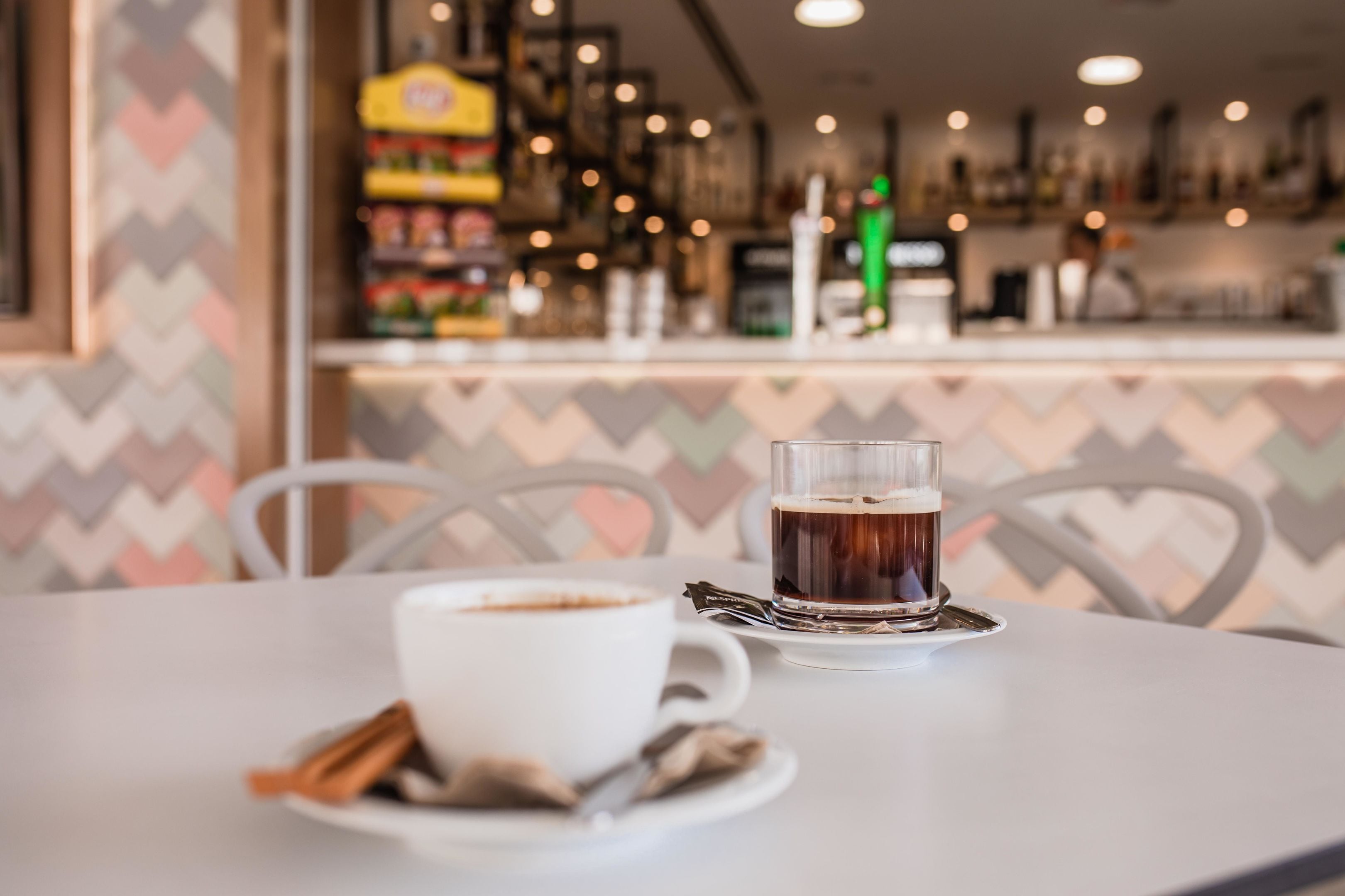 Ein Tisch mit einer Tasse Kaffee und einem Teeglas, einem Zimtstange und einem Teebeutel. Im Hintergrund ist eine Bar mit Regalen voller Flaschen und einer gemusterten Wand zu sehen.