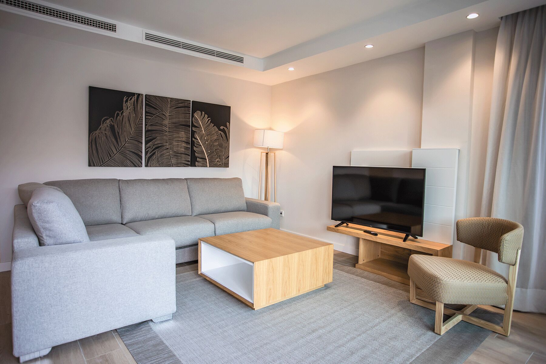 Una sala moderna con un sofá gris, una mesa de centro de madera, un televisor sobre un mueble bajo, una lámpara de pie y una silla estampada.