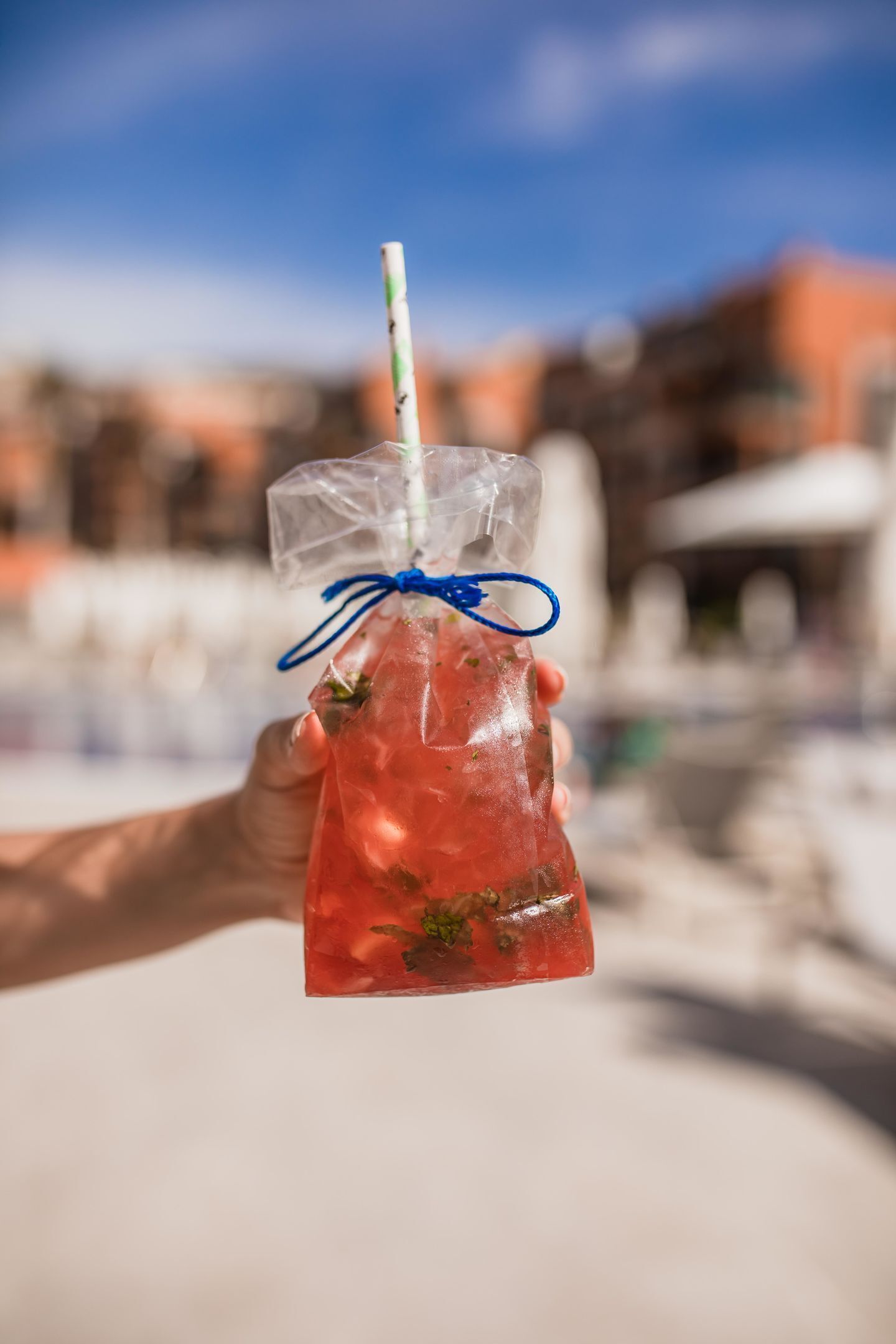 A hand holding a clear plastic bottle with a blue ribbon and a straw, containing a red drink with green herbs, against a blurred background of buildings.