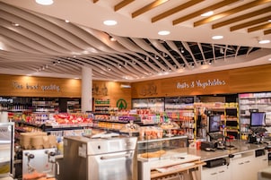 Interior de una tienda de comestibles con estantes llenos de diversos productos, una charcutería y un letrero que indica la sección de frutas y verduras.