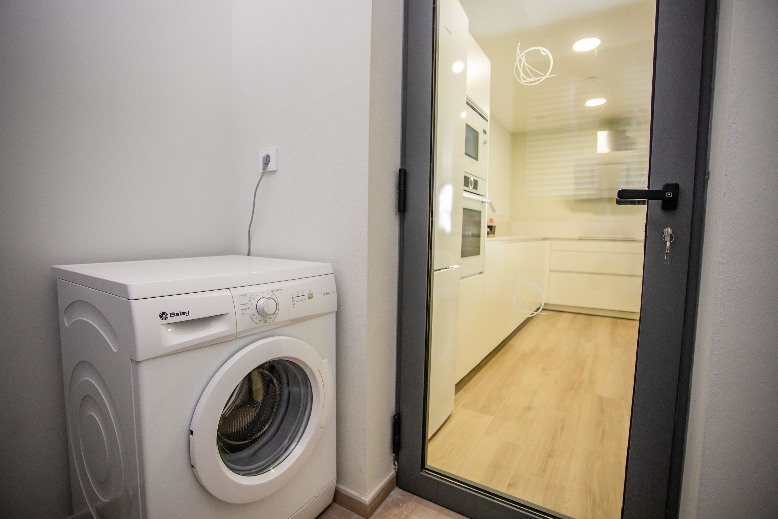 Un lavadero pequeño con lavadora de carga frontal, una puerta que da a una cocina con suelo de madera y una lámpara moderna.
